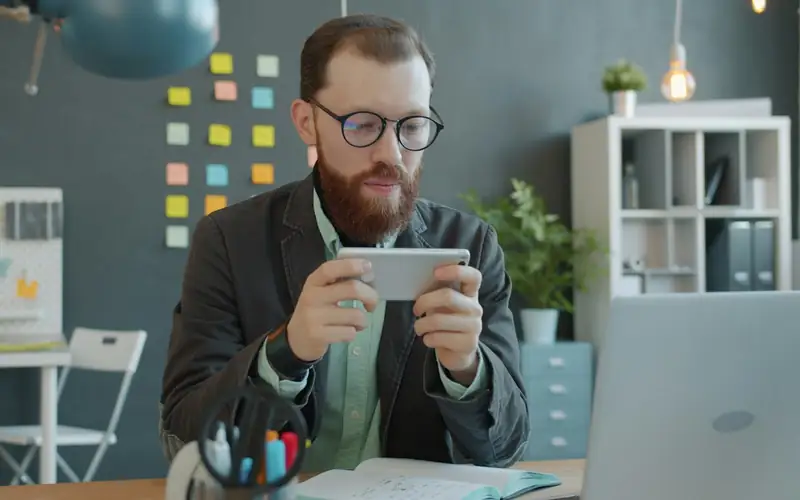 Bearded man holding a smartphone sideways to play a mobile game during a break at his desk